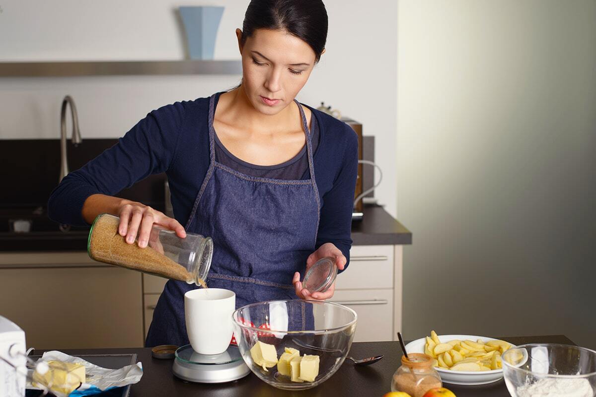 Giovane donna in piedi davanti al bancone della cucina con indosso il grembiule mentre pesa gli ingredienti per aggiungerli alla ciotola contenete il burro